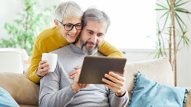 A man reviews a tablet on his sofa while his wife hugs him from behind. 
