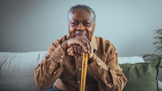 A seated man smiles while resting his chin on his cane handle. 