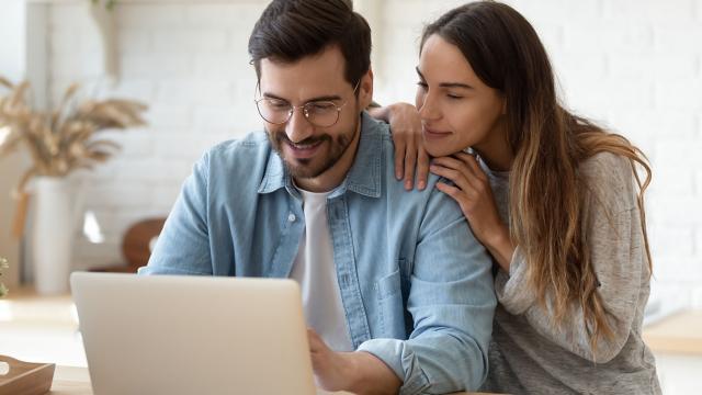 A man uses a laptop while his partner rests her chin on his shoulder. 