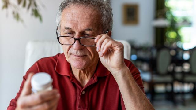 A man wearing glasses studies the fine print on a bottle of pills. 