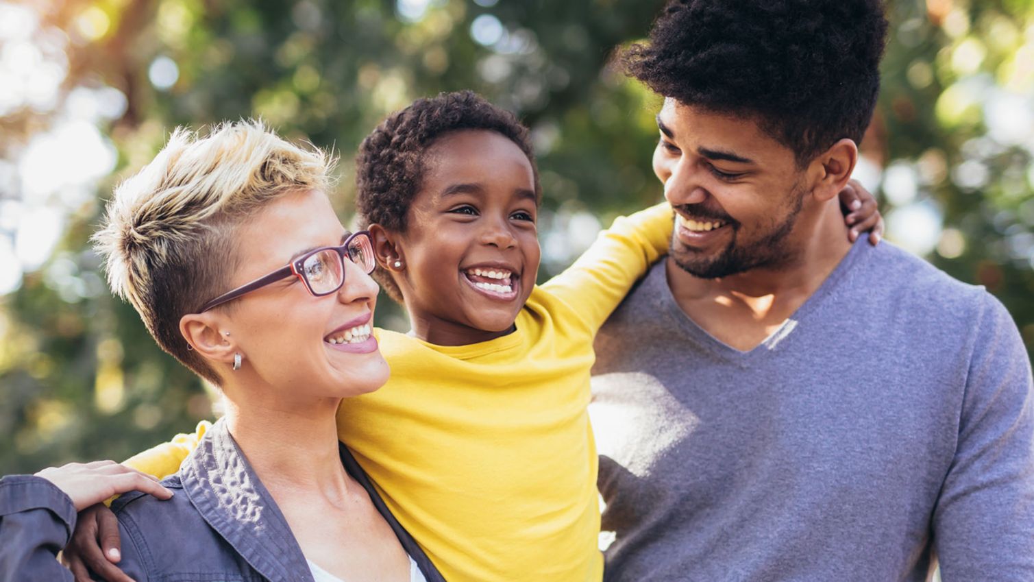 A child grins as she hugs her parents aorund their necks. 