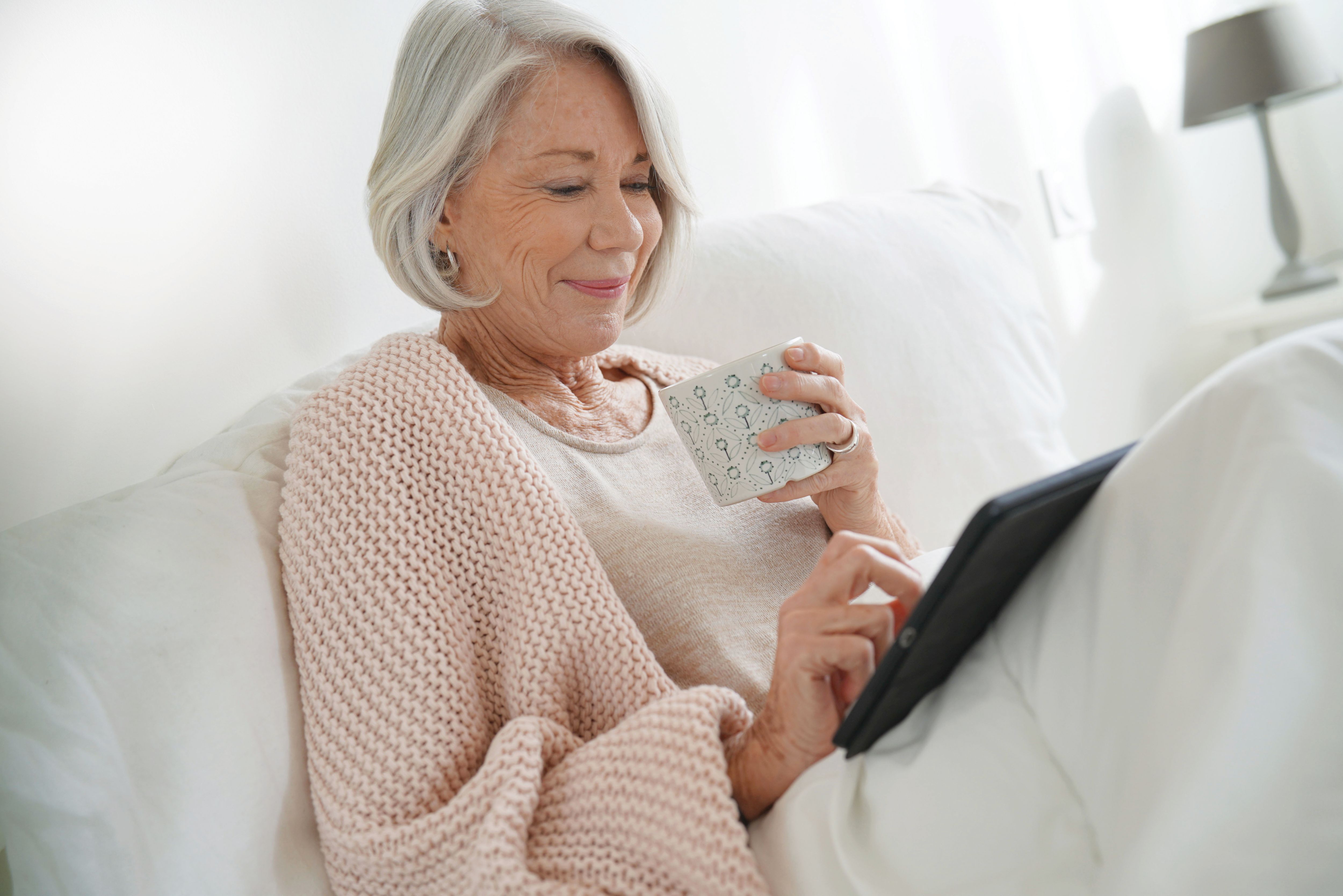 Senior woman relaxing in bed with hot drink and tablet