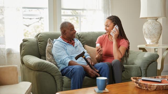 A man sits on his couch with his adult daughter while she uses a telephone.