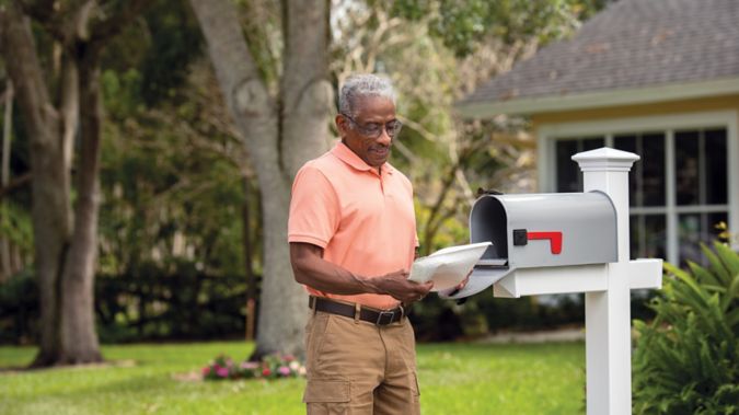 A man stands near his mailbox and smiles at a package he received.