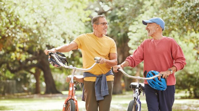 A couple smiles and chats while walking their bikes along a path in a park.