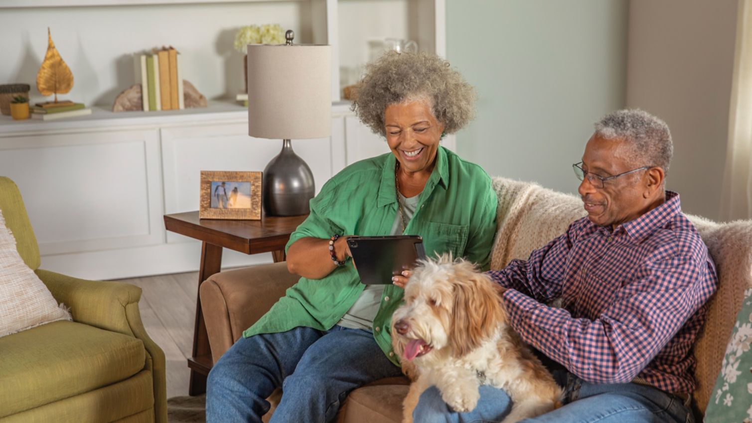 A couple sits on their sofa with their dog while the woman uses a tablet. 