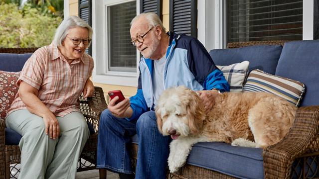 A couple sits with their dog on a porch and smiles while looking at a smartphone.