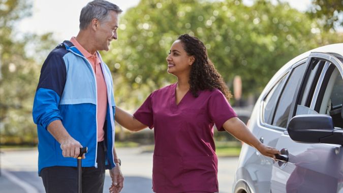 Senior man with cane is assisted by female caregiver walking away from car