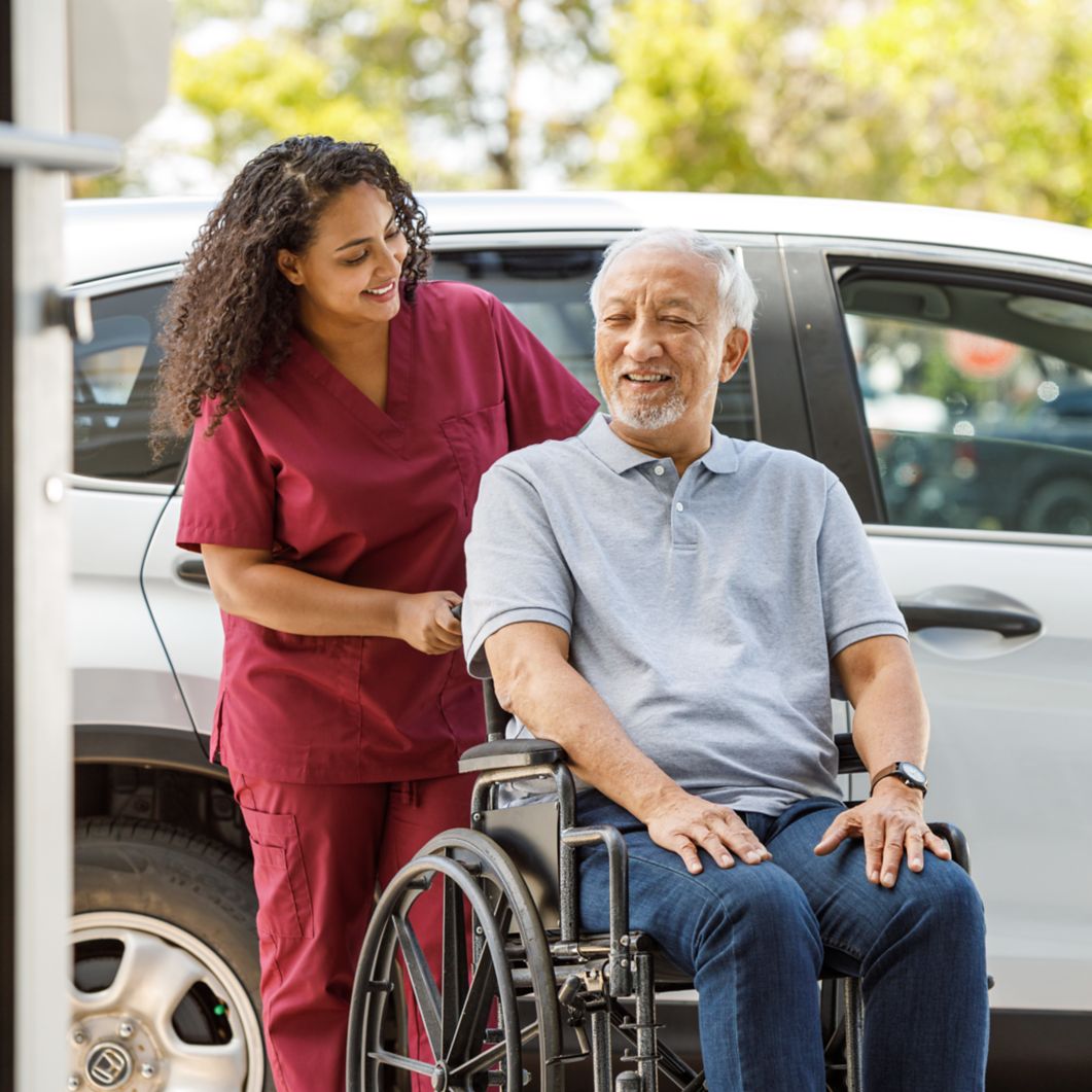 A man in a wheelchair is pushed by a nurse from his car to a building. 