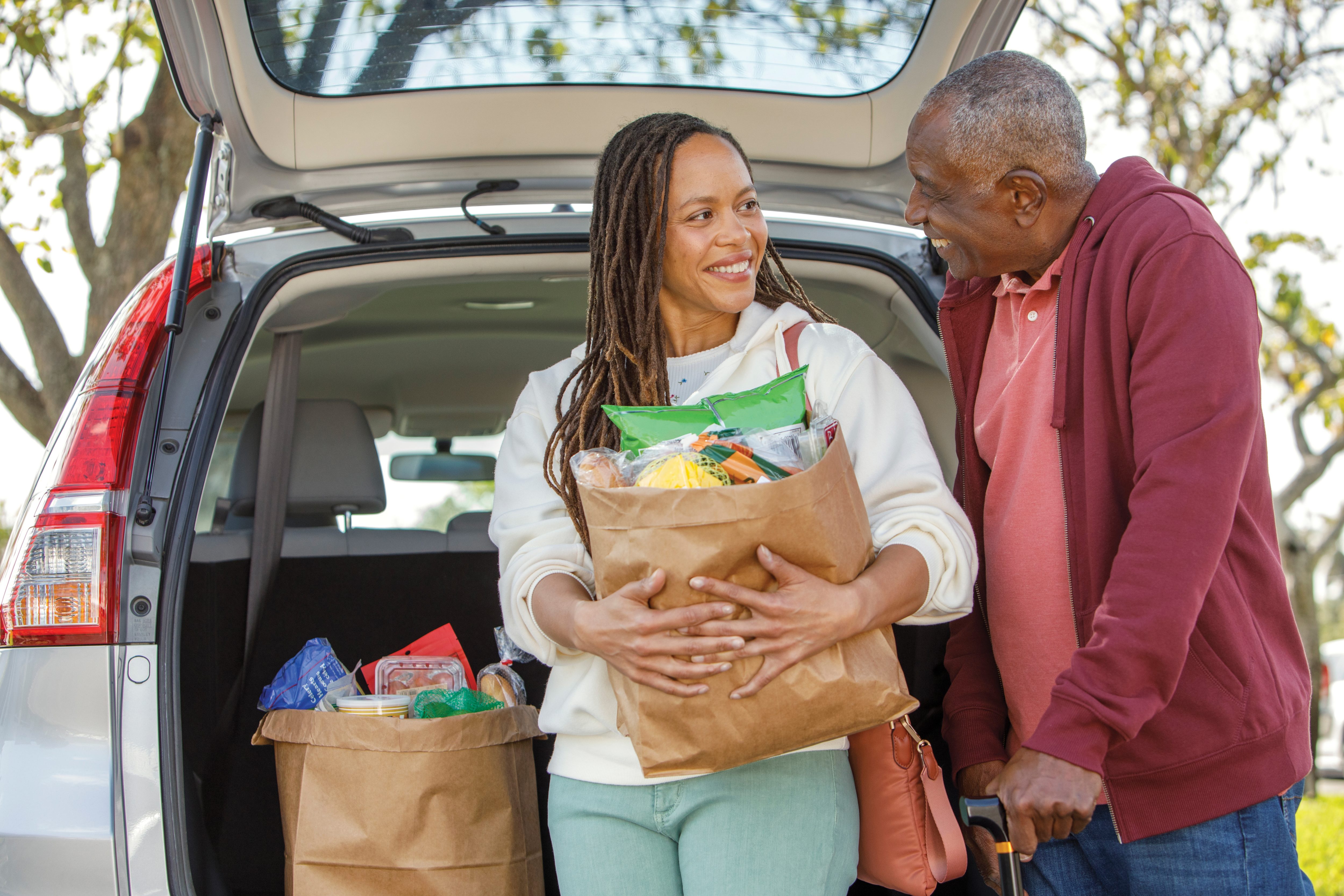 Adult daughter helps unload groceries from car trunk for Senior man with cane