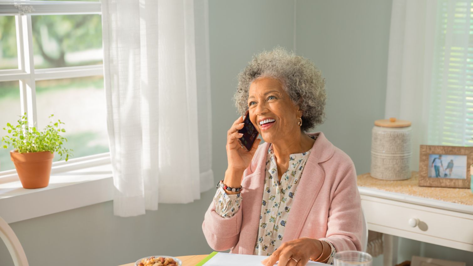 A woman smiles at her kitchen table while making a phone call. 