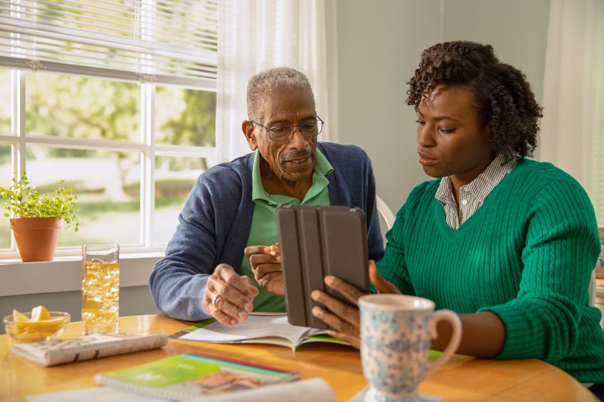 A man looks at a tablet his daughter holds as they sit at his kitchen table.