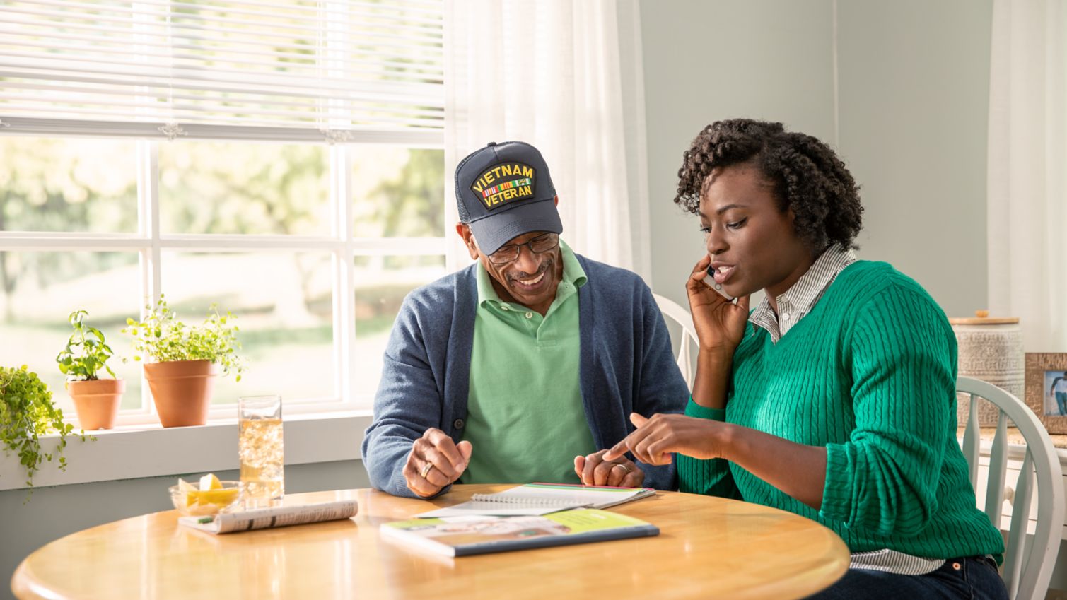 A man smiles at his adult daughter while she makes a phone call at his kitchen table. 