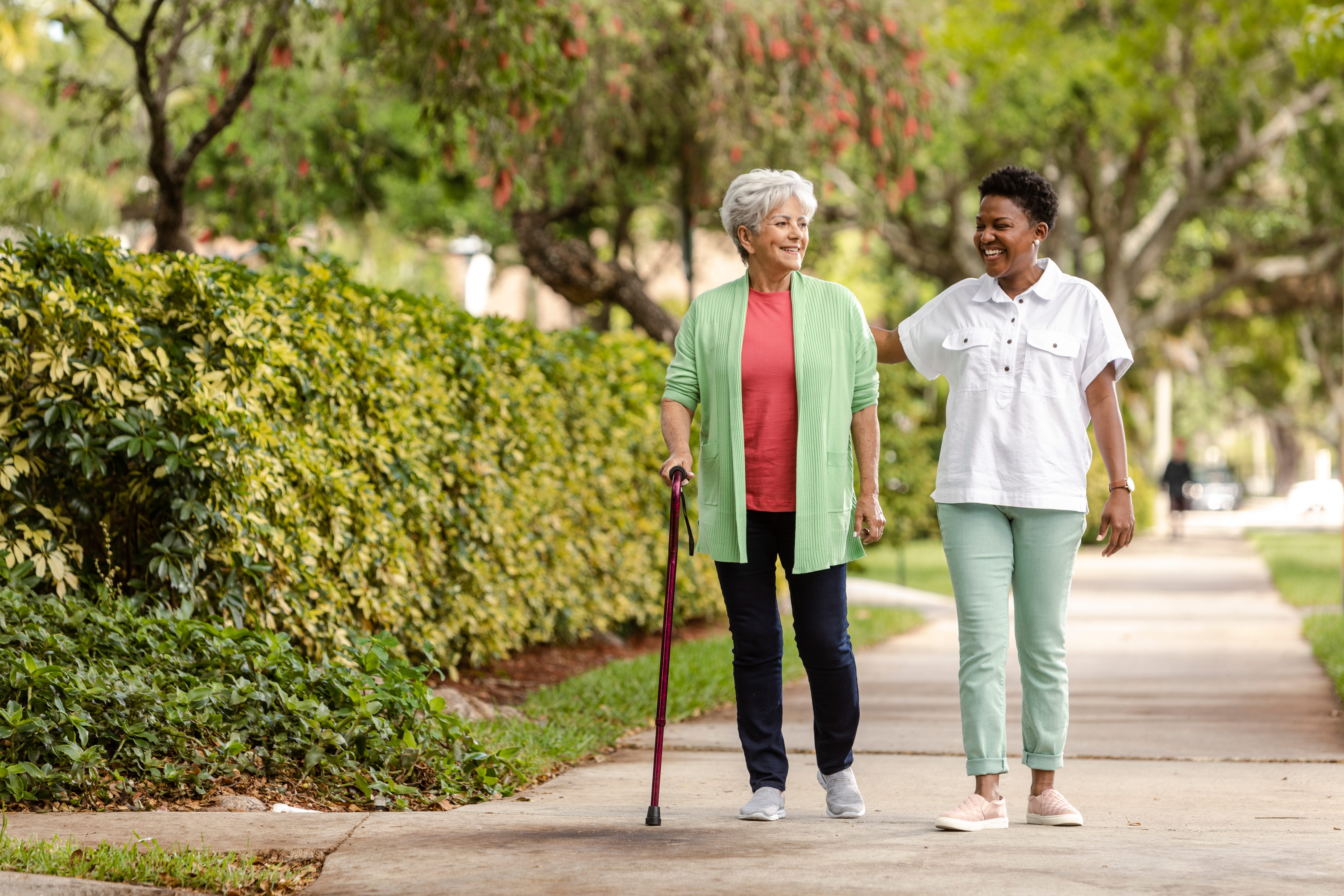 Senior Woman using a cane receives assistance from caregiver as they take a walk outdoors