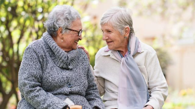 Two ladies lean in for a laugh as they relax side-by-side on a park bench.  