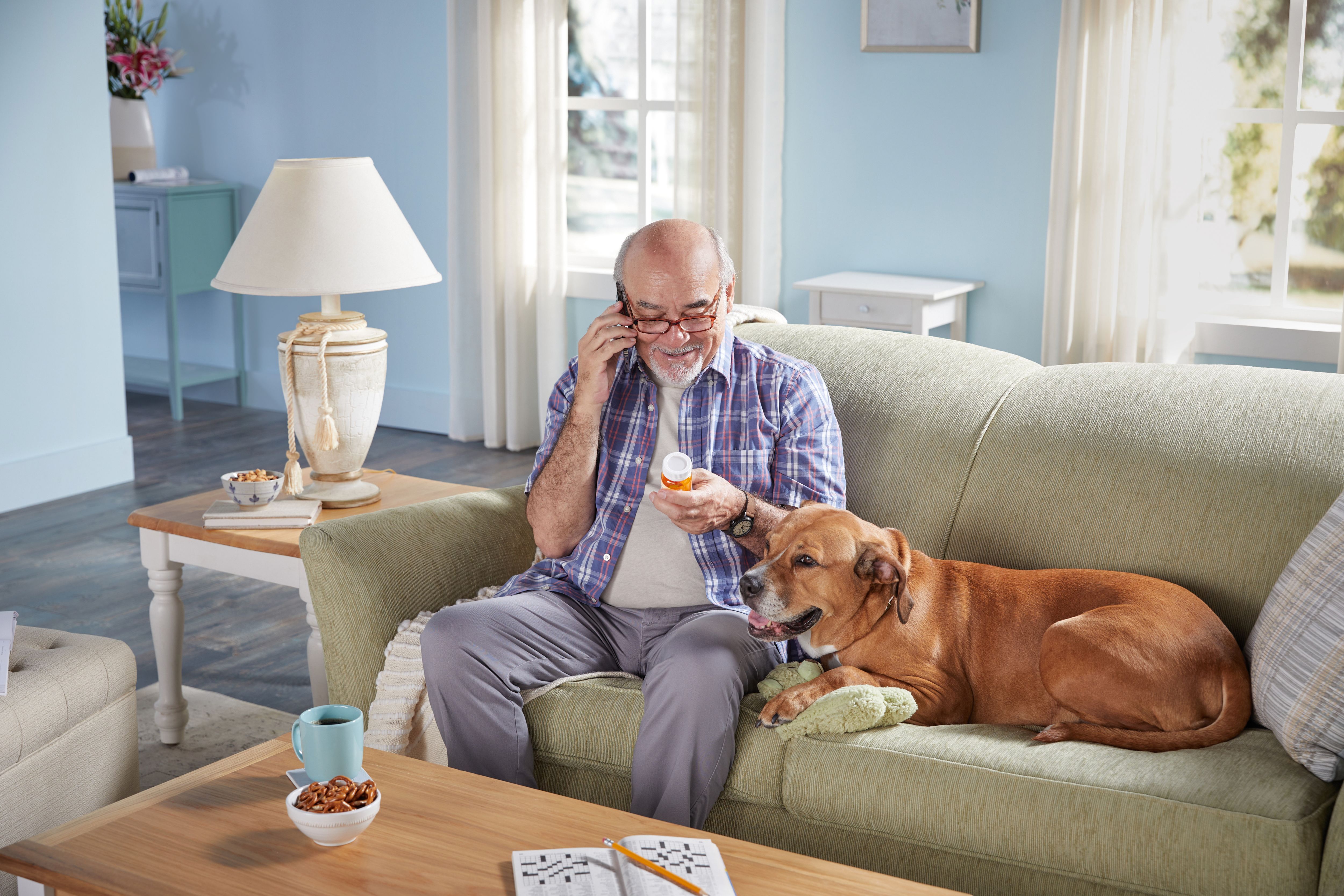 Senior man sitting on couch in living room with his dog reviewing medications while on the phone