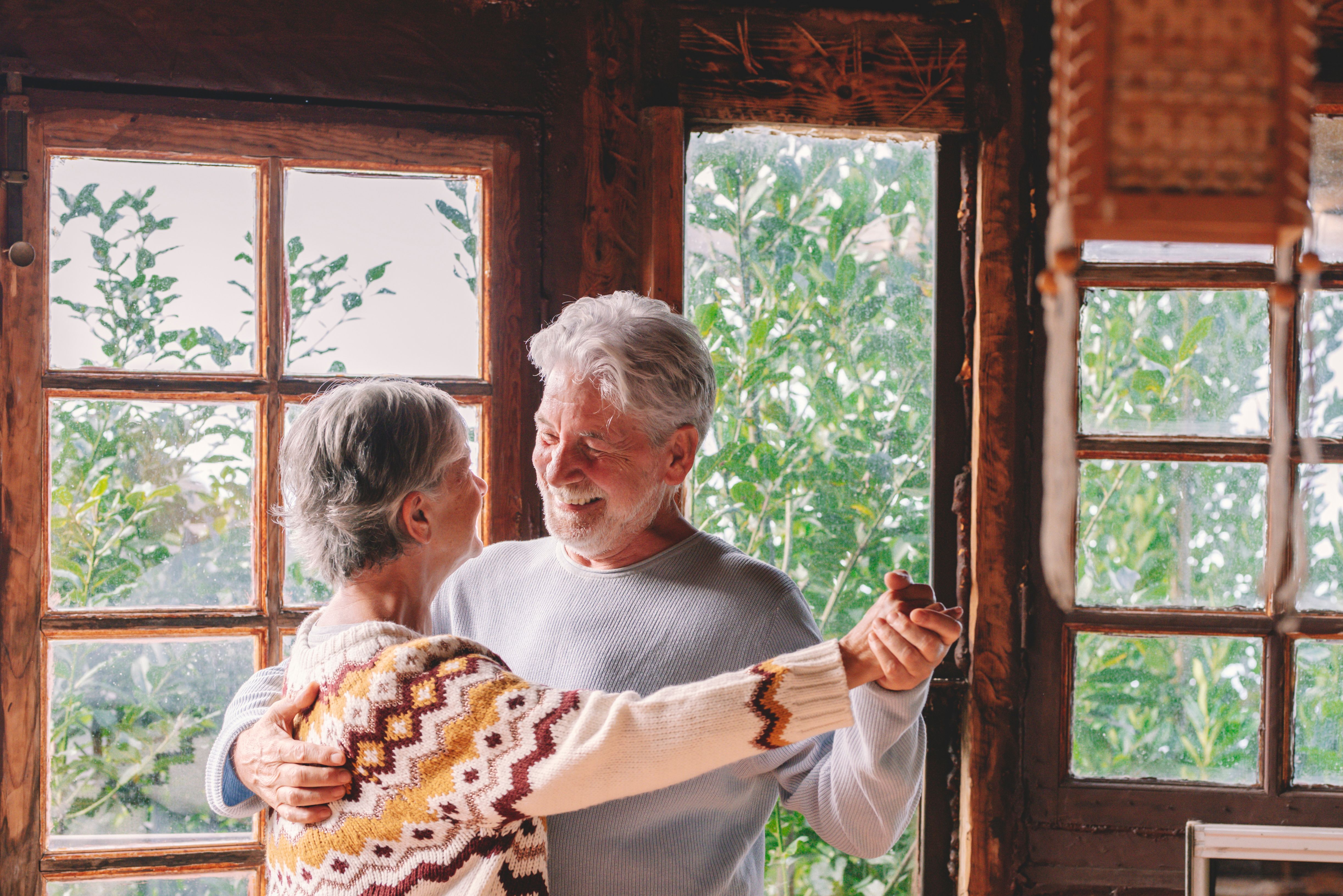 Happy senior couple smile and dance at home enjoying love and relationship together. Active old man and woman have fun in indoor leisure activity. Nature woods view from windows