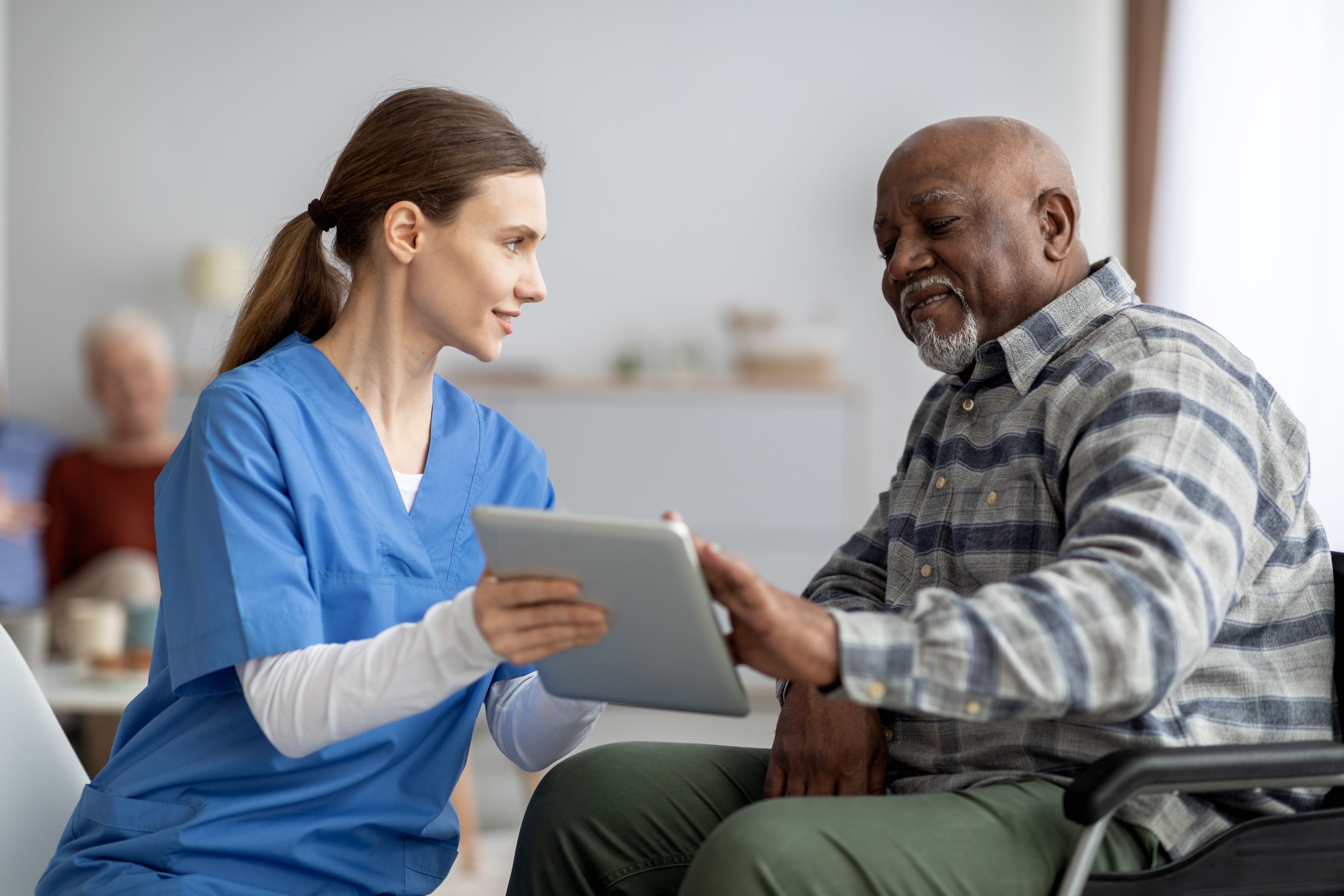 Friendly young lady in workwear nurse helping black senior man in weheelchair using modern digital tablet, elderly african american male patient surfing on Internet at nursing home