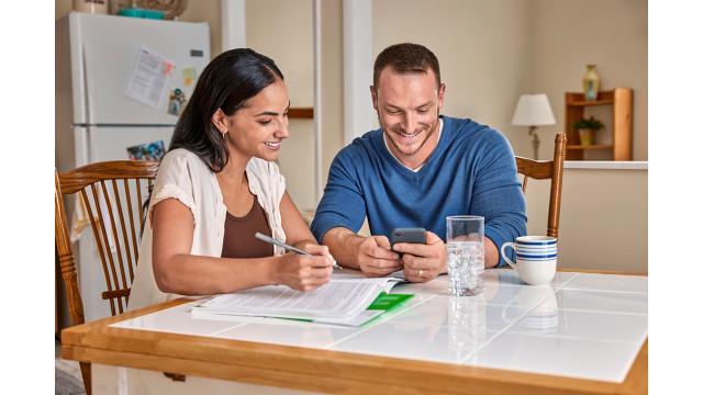 A woman and a man sit at a table, reviewing documents while using a smartphone, a glass of water and a coffee cup are nearby