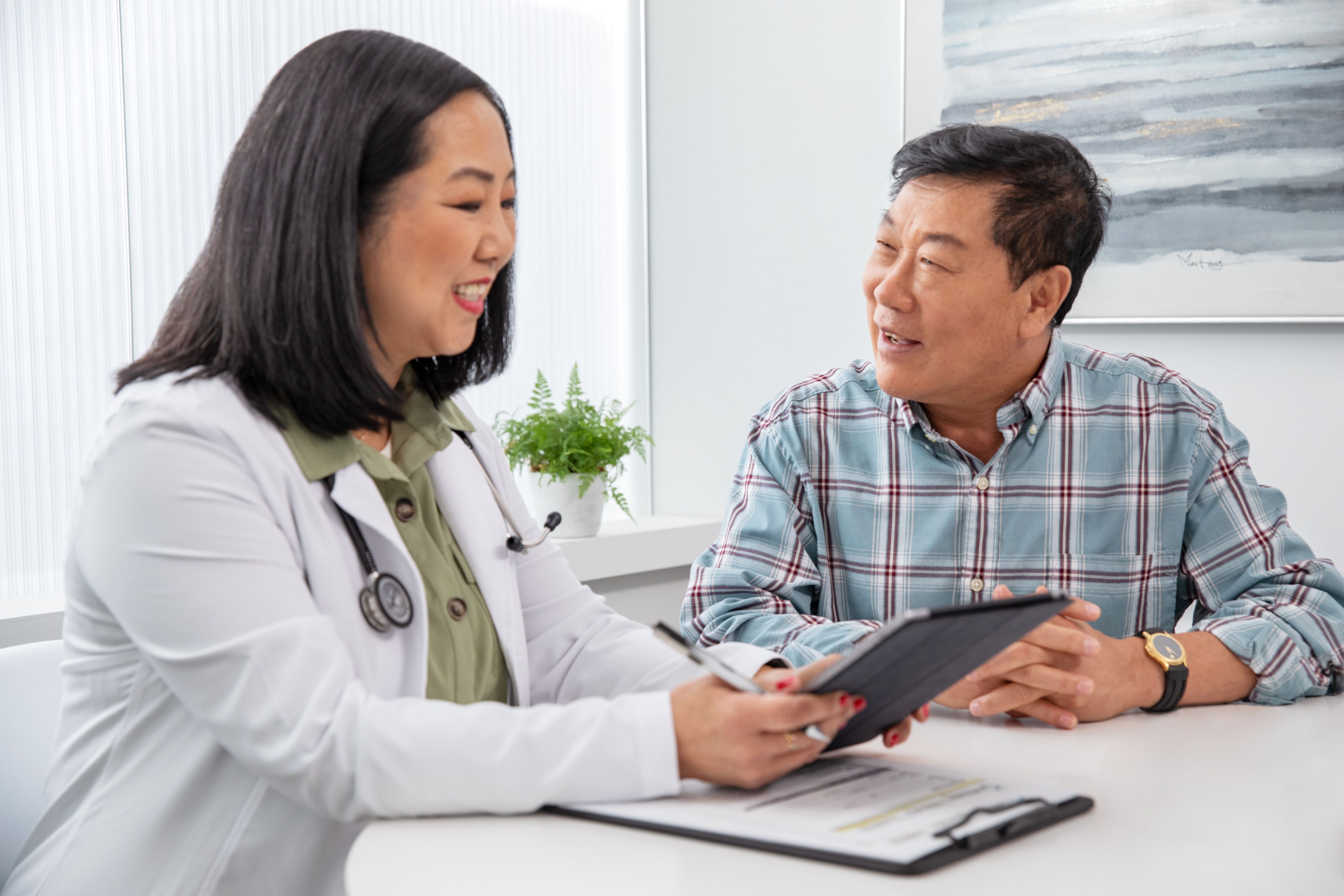 Adult woman doctor sitting at table holding a tablet consulting a senior man 