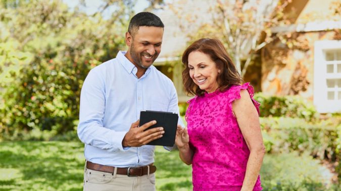 Agent reviewing plan options on tablet wth woman standing in front of house