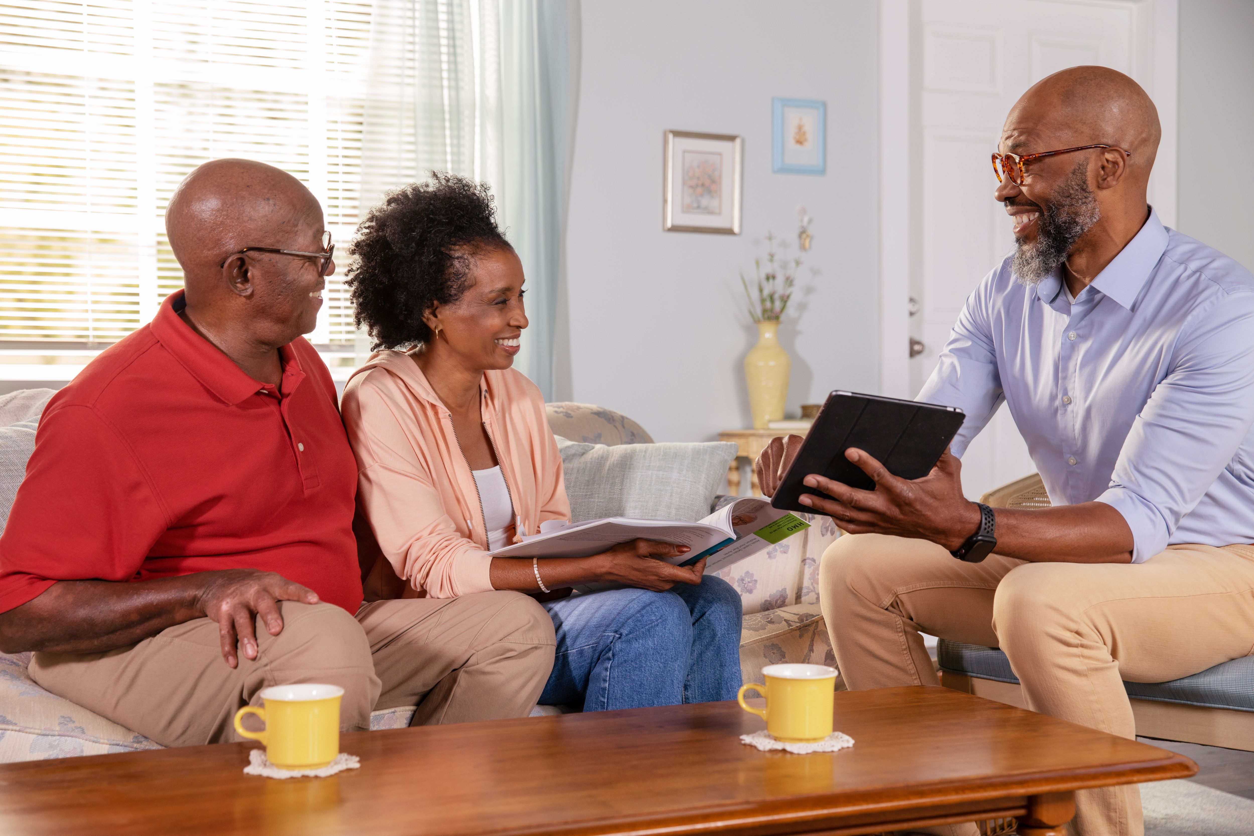 Adult man agent sitting in living room reviewing a tablet with Senior couple smiling on couch.