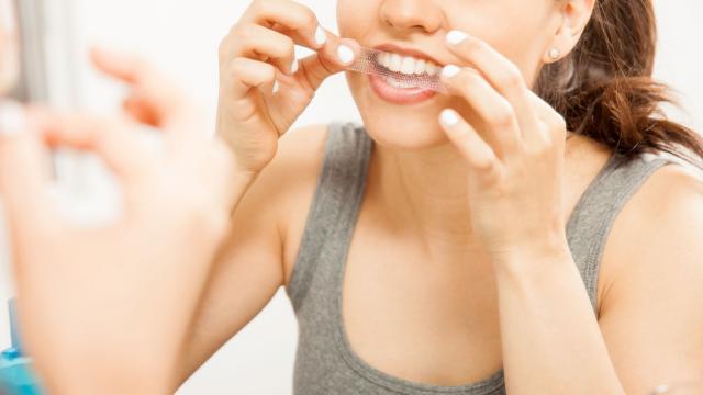 A woman looks in a mirror to apply whitening strips to her teeth. 