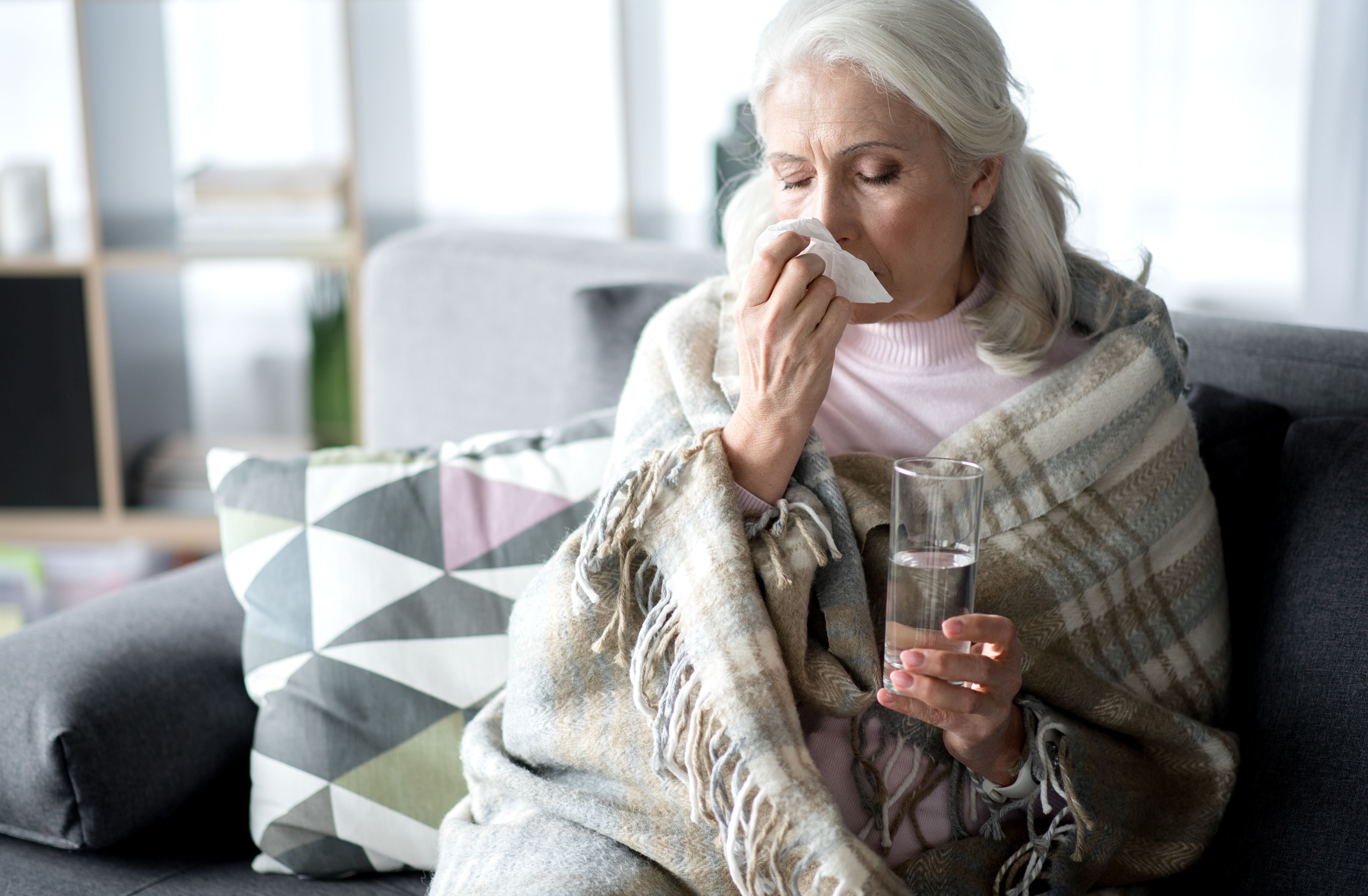 Sick elderly woman is having runny nose. She is wiping it by napkin while sitting on couch and holding glass of water. Copy space