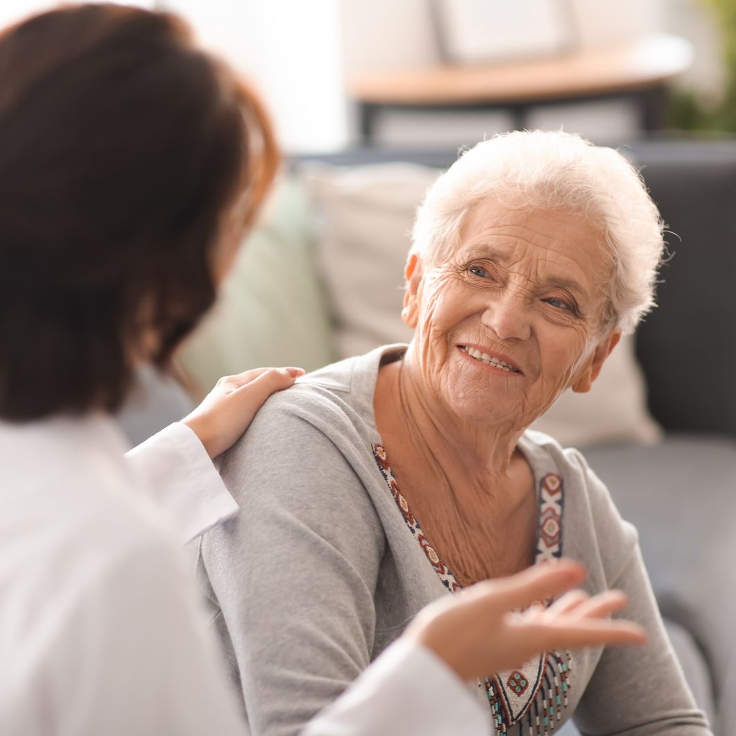 An elderly woman with white hair smiles warmly while a caregiver in a white lab coat places a hand on her shoulder.