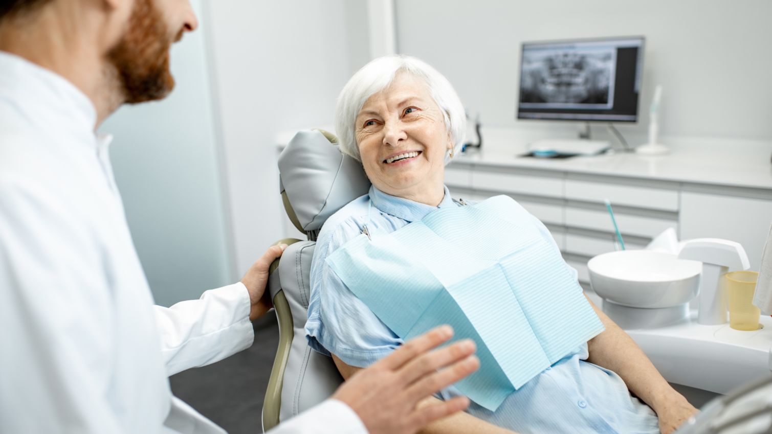 A woman sits in an exam chair and smiles while talking with her dentist. 