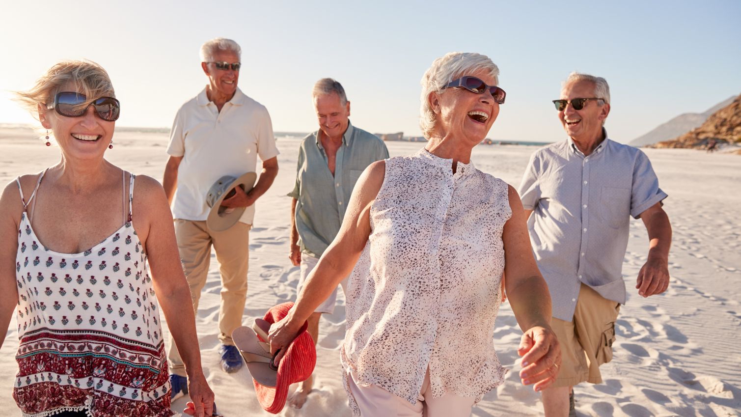 A group of friends laugh as they walk together on a windy beach at sunset.