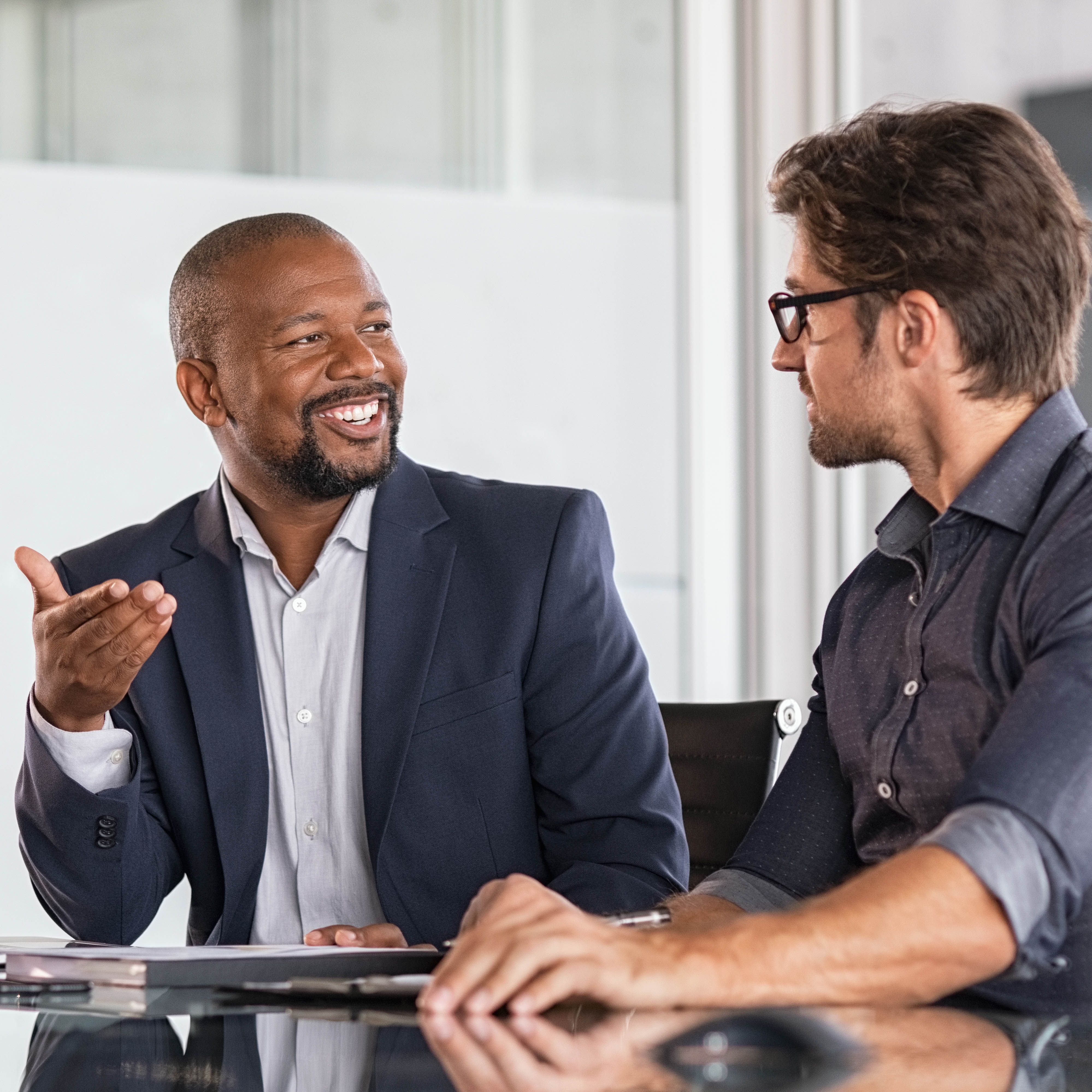 Two happy business colleagues at meeting in modern office interior. Successful african boss in a conversation with young employee in boardroom. Marketing team of two businessmen discussing strategy in meeting room.