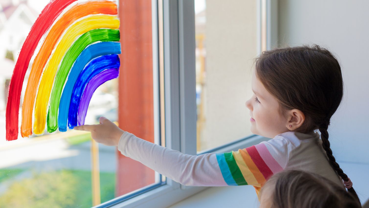 little boy at home behind a window holding up a rainbow drawing