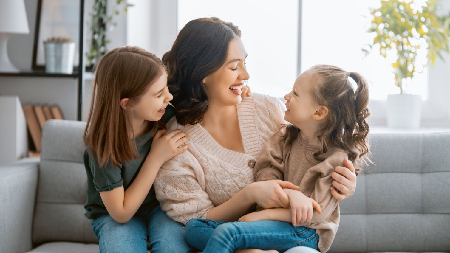 A mom laughs with her young daughters on their sofa. 