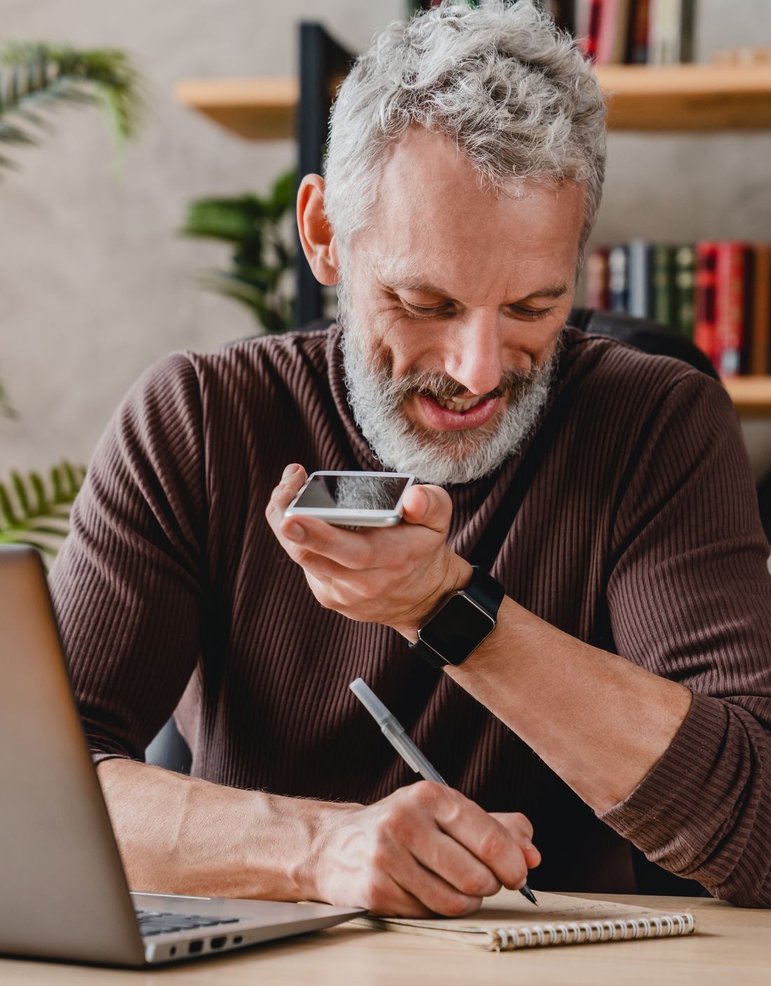 Senior man writing notes in notepad while talking by speakerphone from workspace