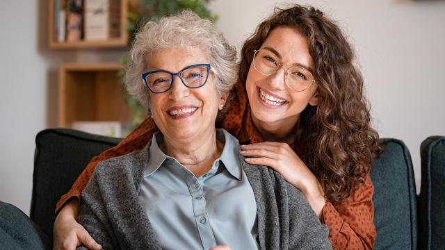 A woman smiles while her adult daughter hugs her from behind. 