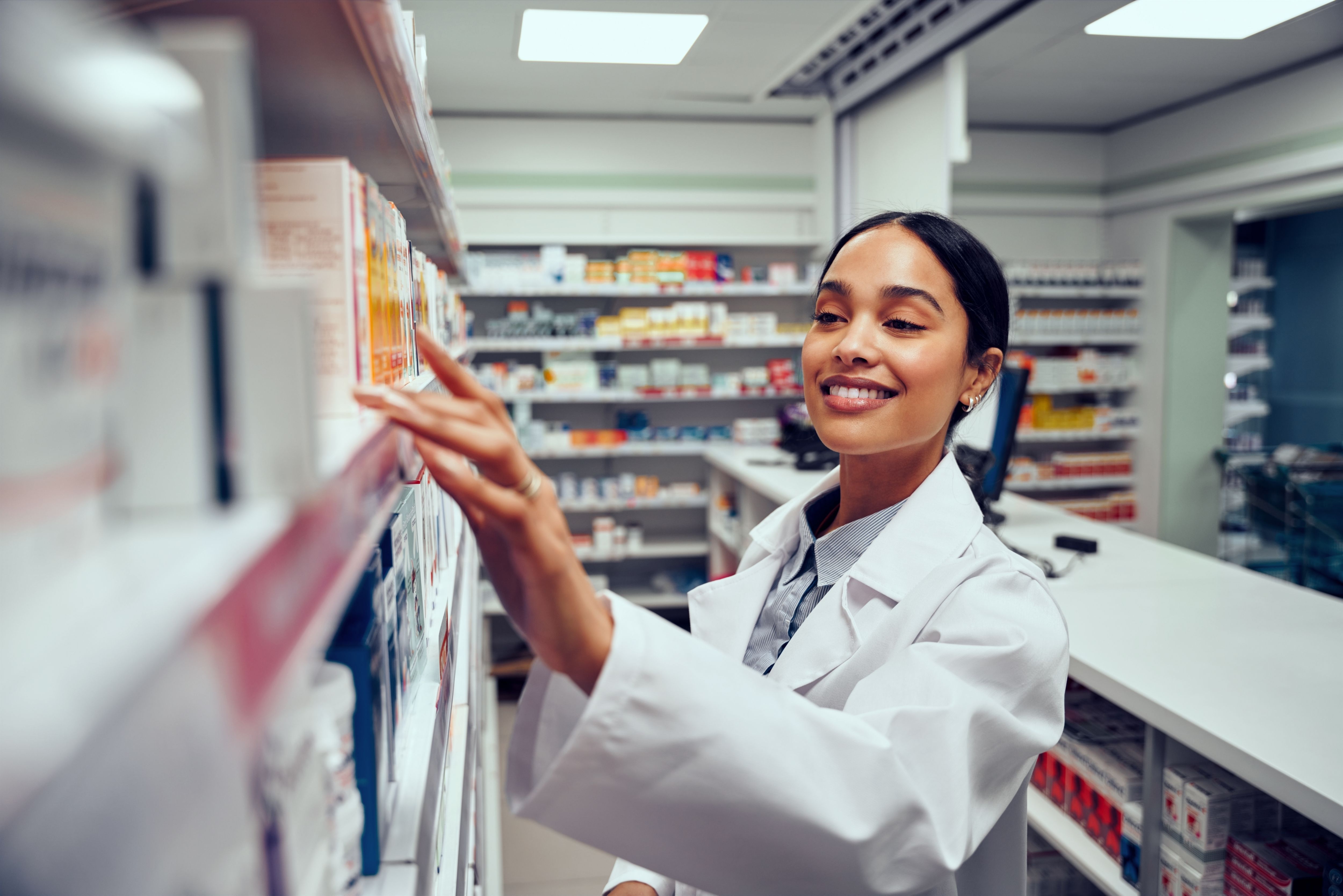 Smiling young female pharmacist behind counter looking for medicine in shelf
