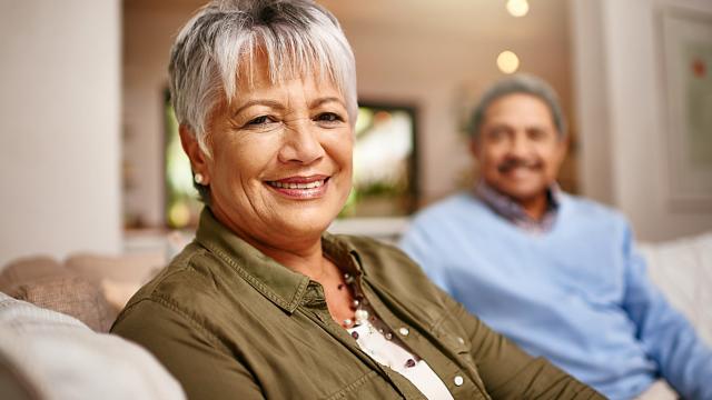 A woman on her sofa smiles while her husband looks on from the background.