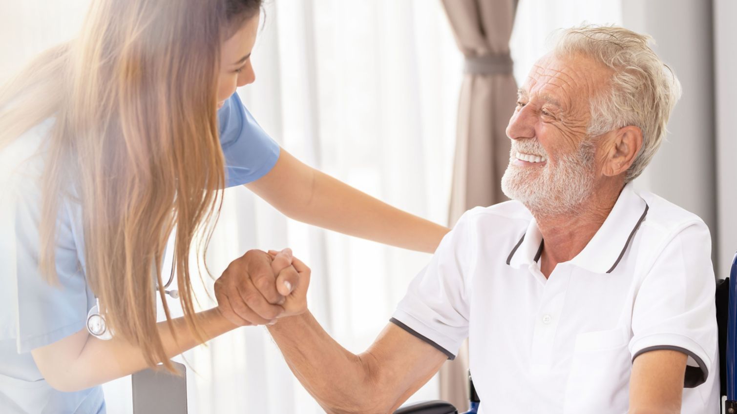 A young female caregiver clasps hands and smiles with an elderly man seated in a wheelchair.