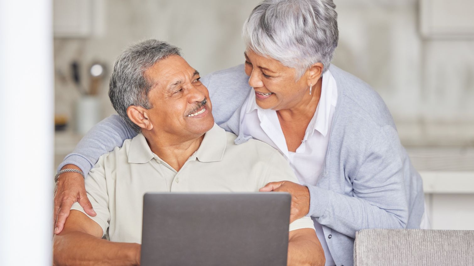 Man sitting at his laptop turns to smile as his wife who has snuck up with a hug. 