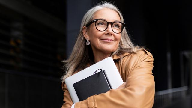 A woman smiles while holding a laptop and a notebook. 