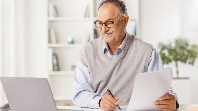 A man takes notes while reading from his laptop screen. 