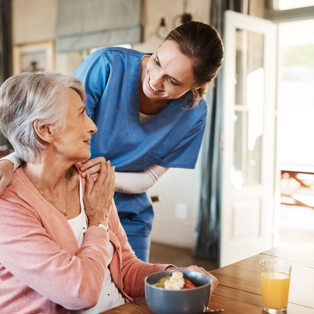 A healthcare worker stands over an elderly woman with her arm around her shoulder