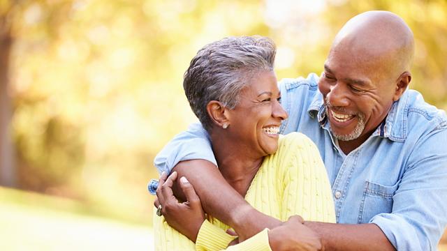 A man hugs his wife from behind while they grin at each other. 