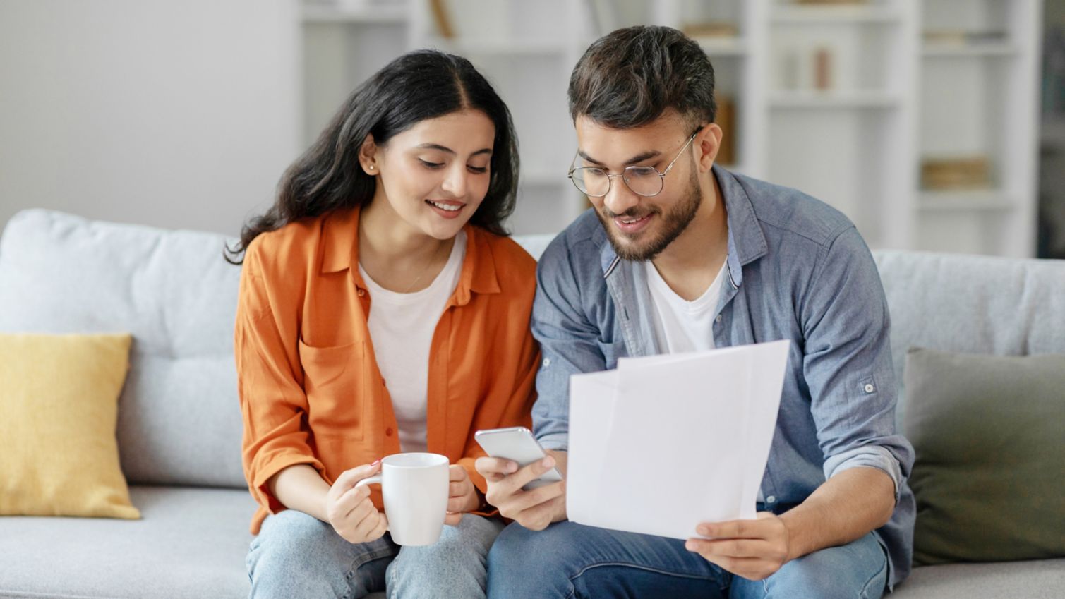 Spouses sitting on sofa, examining document together with cellphone 
