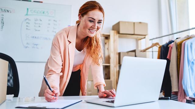 Smiling woman takes notes while reading from her laptop.