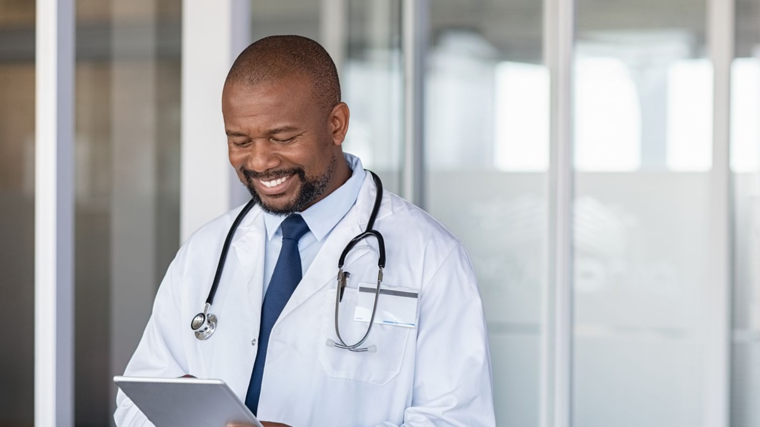 A male physician stands in a hallway smiling at his tablet