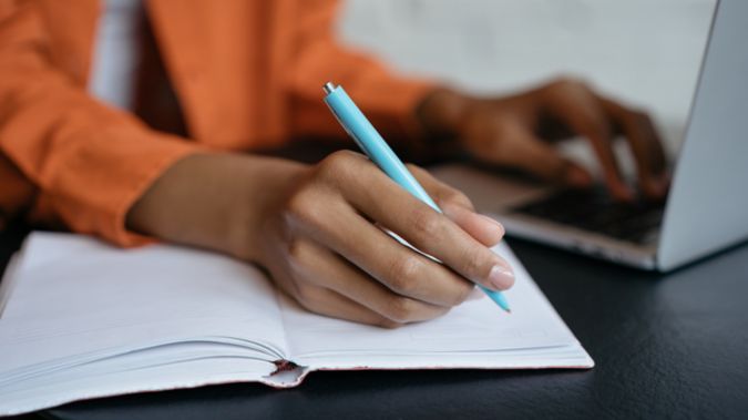 A woman writes in her notebook while working at a computer