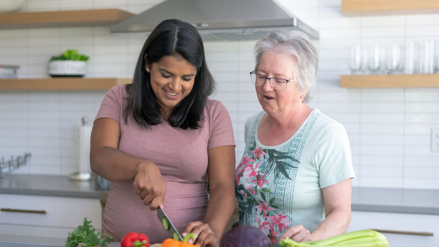 Pregnant woman chops vegetables on kitchen counter as older woman looks on