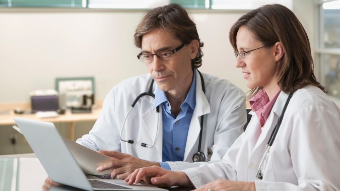 Two physicians sit at a table and discuss information on a laptop screen